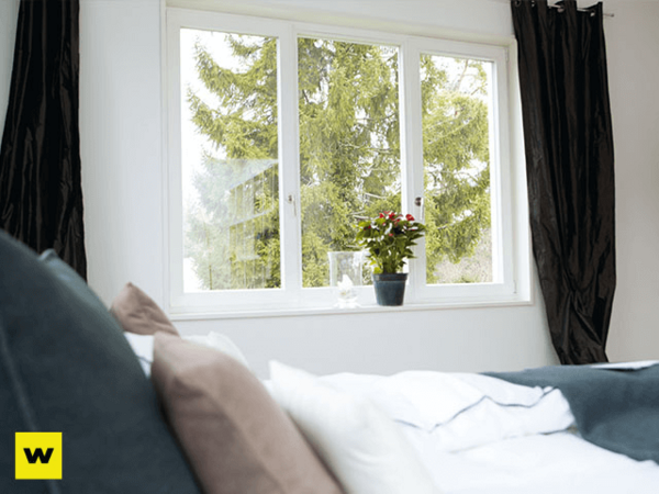 Helles Schlafzimmer mit Bett, großen Fenstern, Vorhängen und Topfpflanze auf der Fensterbank.