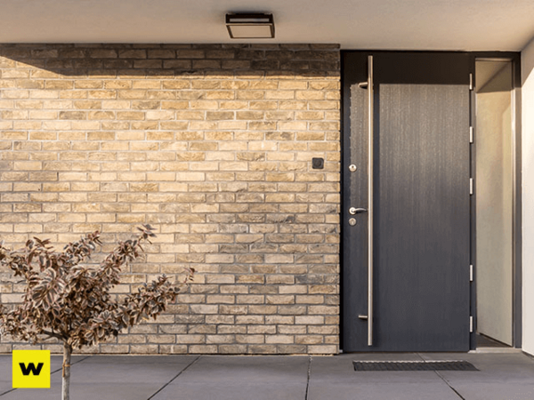 Moderne Haustür aus dunklem Material in einer Fassade mit Klinkerstein und kleinem Baum.